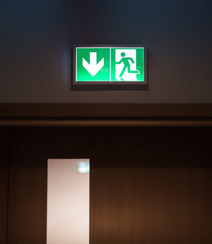 illuminated-emergency-exit-sign-above-doorway-29568751 Green emergency exit sign with arrow above a closed door for evacuation purposes.
