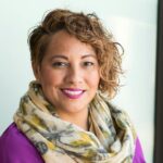 Smiling woman in a purple dress and scarf holding a tablet indoors.